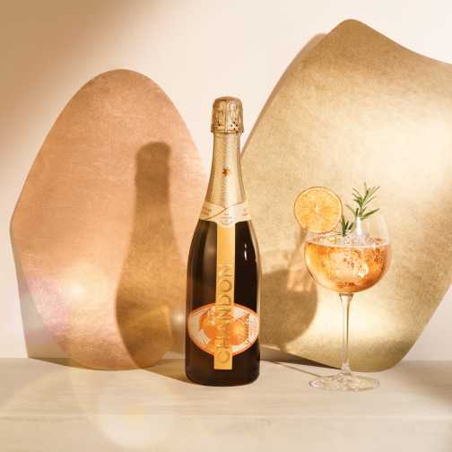 A bottle of CHANDON Garden Spritz and a glass of the spritz cocktail garnished with a dried orange slice and rosemary, against a modern, minimalist background of two abstract, textured panels in rose gold and gold hues.