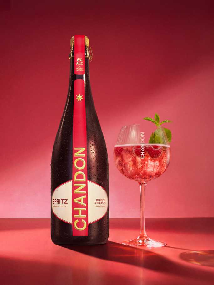 Une bouteille de Chandon Spritz Baies & Hibiscus à côté d'un verre à vin, avec des glaçons, des framboises et des feuilles de menthe dispersés sur un fond rouge vif.