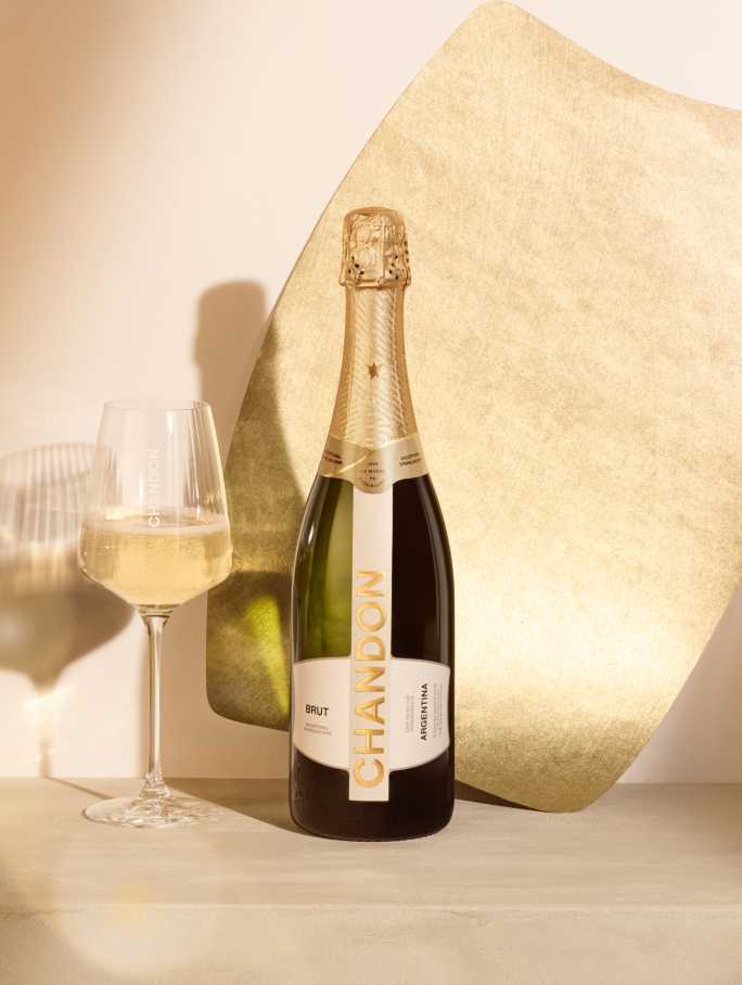 A bottle of CHANDON Brut sparkling wine from Argentina, with a gold and white label, next to an elegant wine glass filled with clear, sparkling white wine, arranged against a modern, minimalist background of an abstract, textured gold panel.