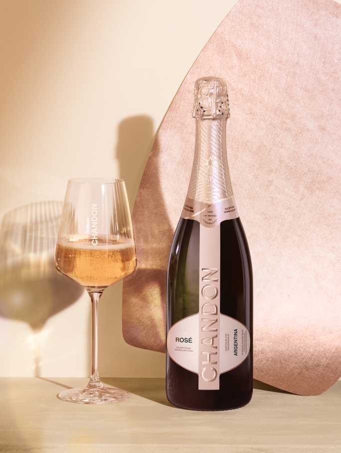 A bottle of CHANDON Rosé sparkling wine from Argentina, with a rose-colored label, next to an elegant wine glass filled with pink sparkling wine, arranged against a modern, minimalist background of an abstract, textured rose gold panel.