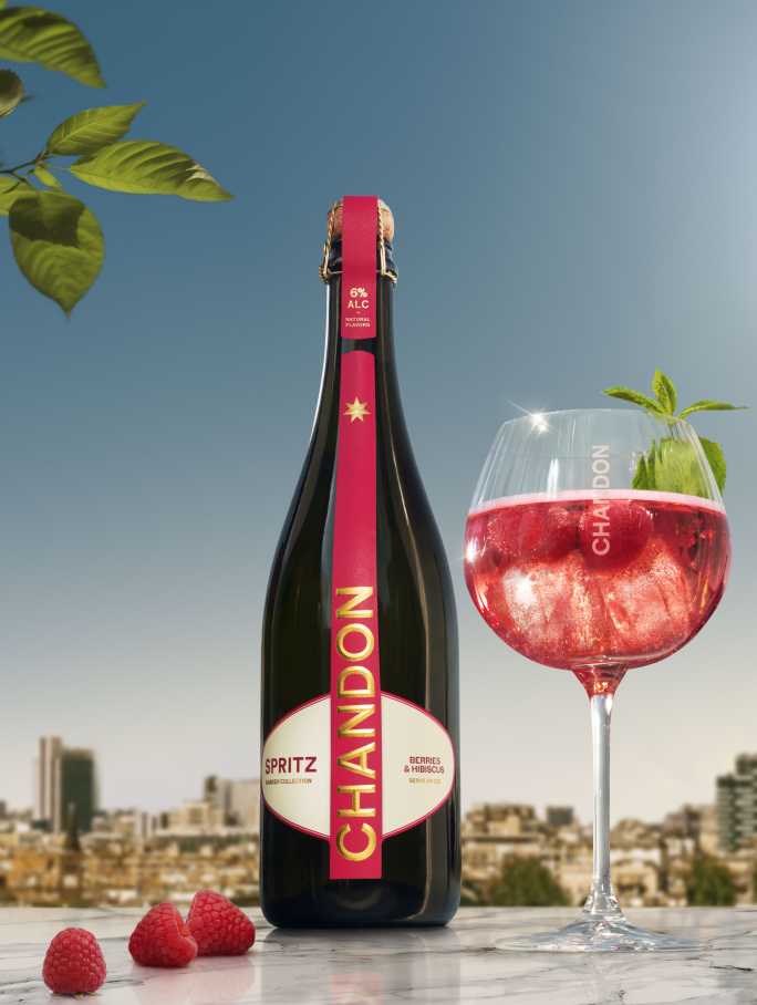 Eine Flasche Chandon Spritz Berries & Hibiscus neben einem Glas Chandon Spritz garniert mit frischen Himbeeren und einem Minzblatt, vor dem Hintergrund einer sonnigen Stadtlandschaft und unter blauem Himmel.
