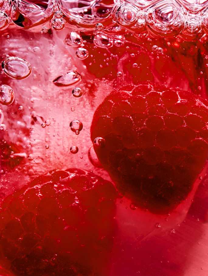 Nahaufnahme von zwei frischen Himbeeren, die in einer sprudelnden, roten Flüssigkeit versinken.
