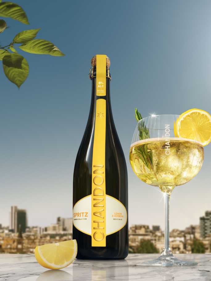A bottle of Chandon Spritz Lemon & Verbena next to a glass of Chandon Spritz cocktail garnished with a lemon slice and a sprig of rosemary, set against a sunny cityscape background.
