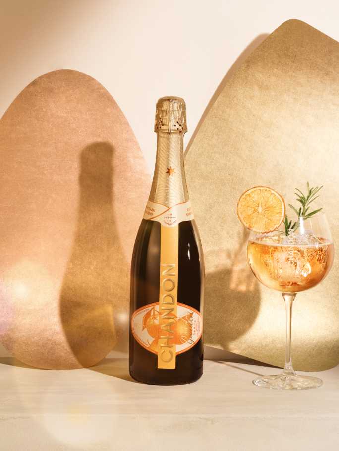 A bottle of CHANDON Garden Spritz and a glass of the spritz cocktail garnished with a dried orange slice and rosemary, against a modern, minimalist background of two abstract, textured panels in rose gold and gold hues.