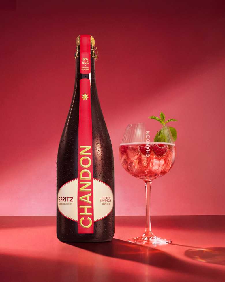 Une bouteille de Chandon Spritz Baies & Hibiscus à côté d'un verre à vin, avec des glaçons, des framboises et des feuilles de menthe dispersés sur un fond rouge vif.