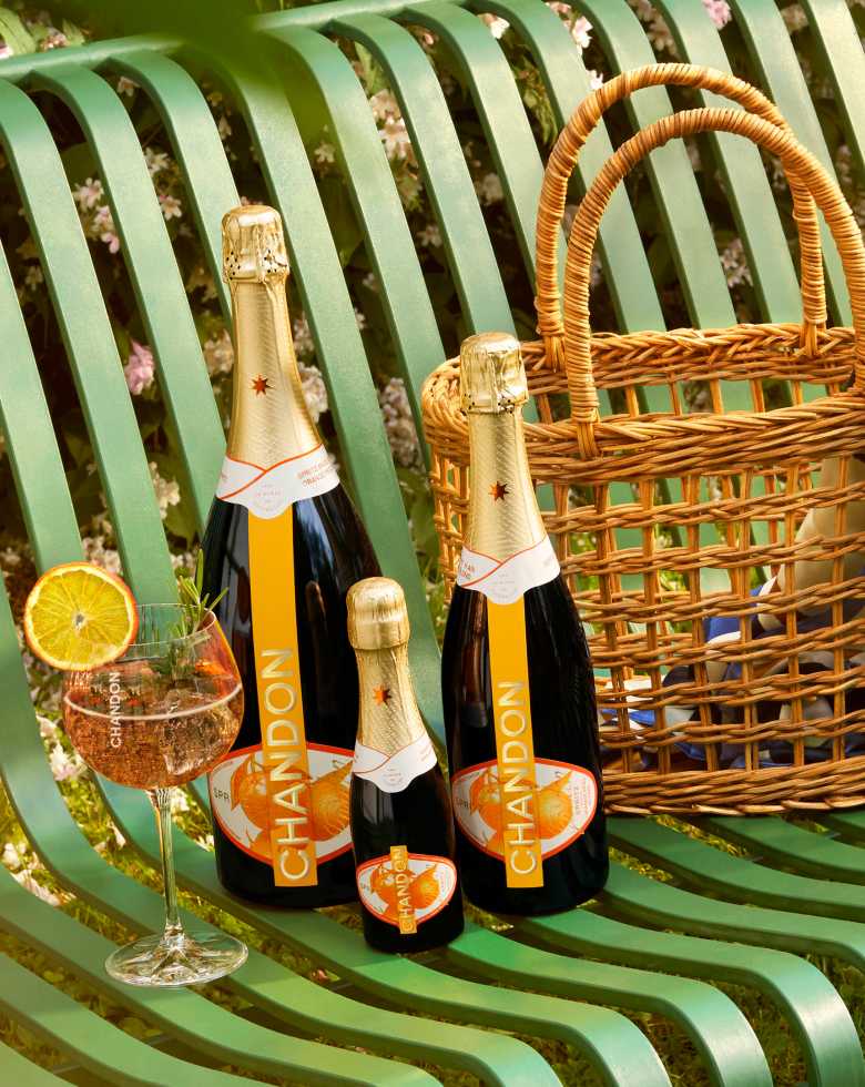 Two sizes of CHANDON Garden Spritz bottles and a small CHANDON mini bottle, arranged with a cocktail glass garnished with an orange slice and rosemary, and a wicker picnic basket with a striped scarf, all resting on a green slatted park bench amidst blooming lilacs.