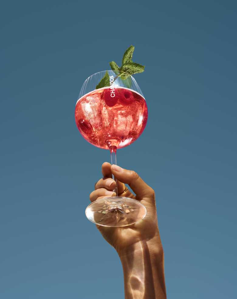 Eine Hand hält ein großes Weinglas mit einem roten Chandon Cocktail in die Höhe, garniert mit frischen Himbeeren und Minzblättern, vor einem klaren blauen Himmel.