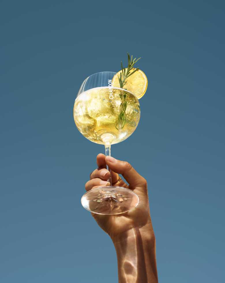 Eine Hand hält ein großes Weinglas mit einem Chandon-Cocktail hoch, garniert mit einer Zitronenscheibe und einem Zweig Rosmarin, vor einem klaren blauen Himmel.