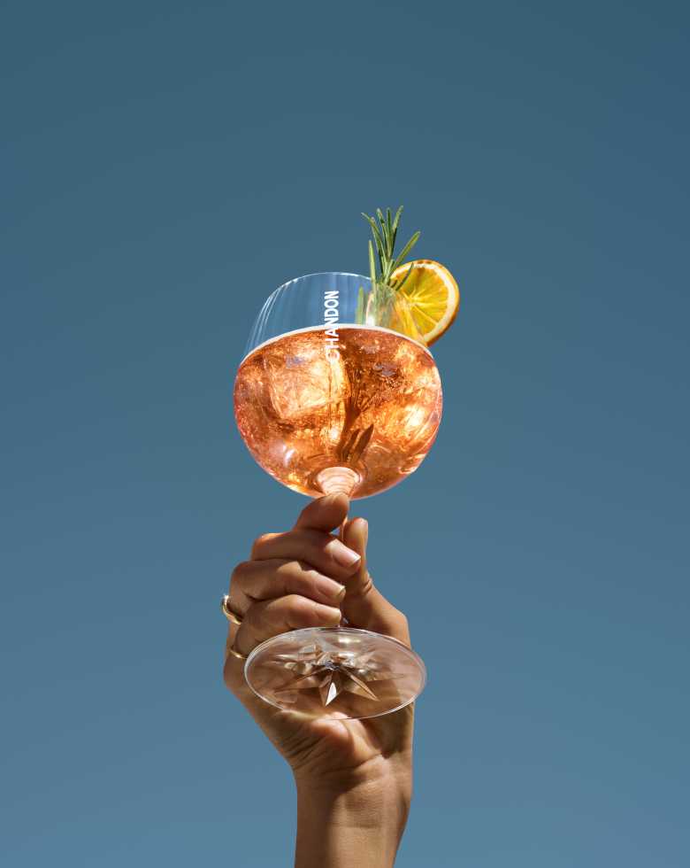 A hand holding up a large wine glass filled with a Chandon Spritz Orange Peels & Spices cocktail, garnished with a dried orange slice and a sprig of rosemary, set against a clear blue sky.