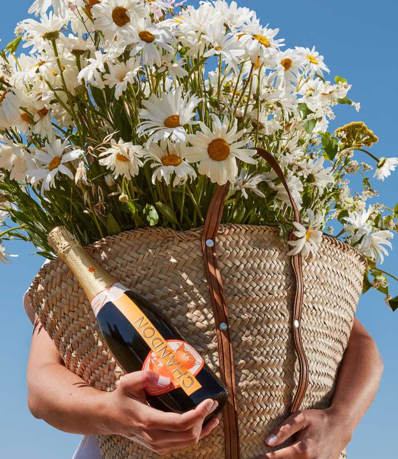 A person holding a woven straw tote bag overflowing with a large bouquet of white daisies, with a bottle of CHANDON Garden Spritz nestled inside the bag, against a clear blue sky.