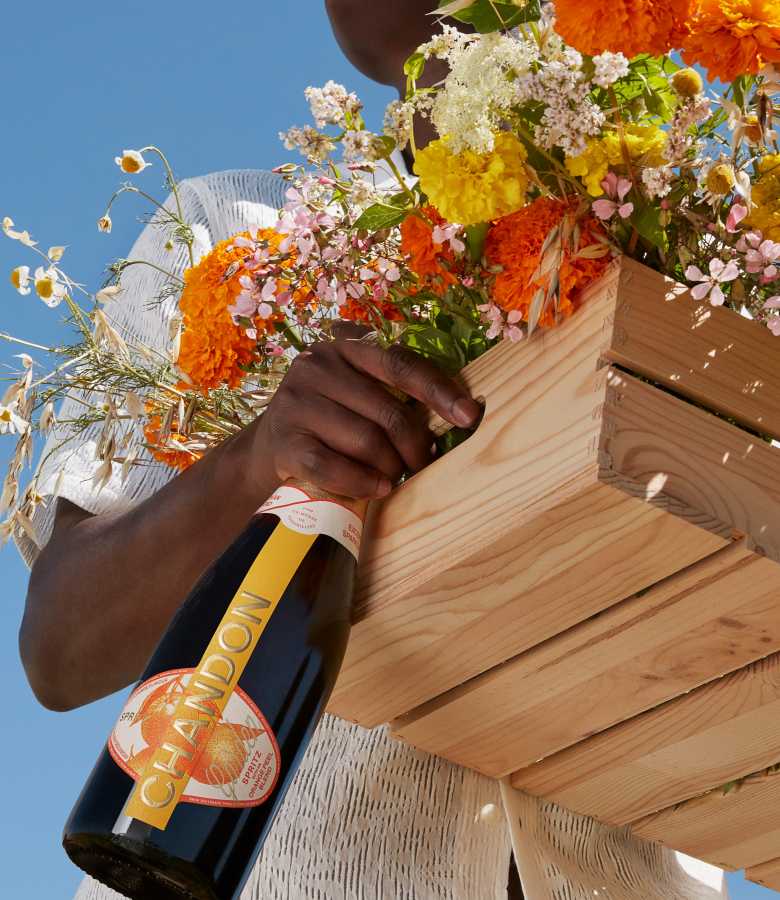  A person holding a bottle of CHANDON Garden Spritz, looking up at the sky, with a rustic wooden crate filled with colorful flowers like orange marigolds and white daisies, suggesting a natural and artisanal theme.