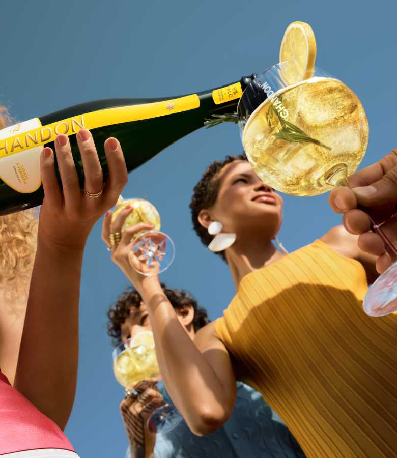 Eine Frau in einem gelben Oberteil und großen weißen Ohrringen lächelt nach oben, während im Vordergrund eine Flasche Chandon Spritz Lemon & Verbena in ein Glas eingeschenkt wird. im Hintergrund ist eine weitere Person zu sehen, die ein Glas hält – alles unter einem klaren blauen Himmel.