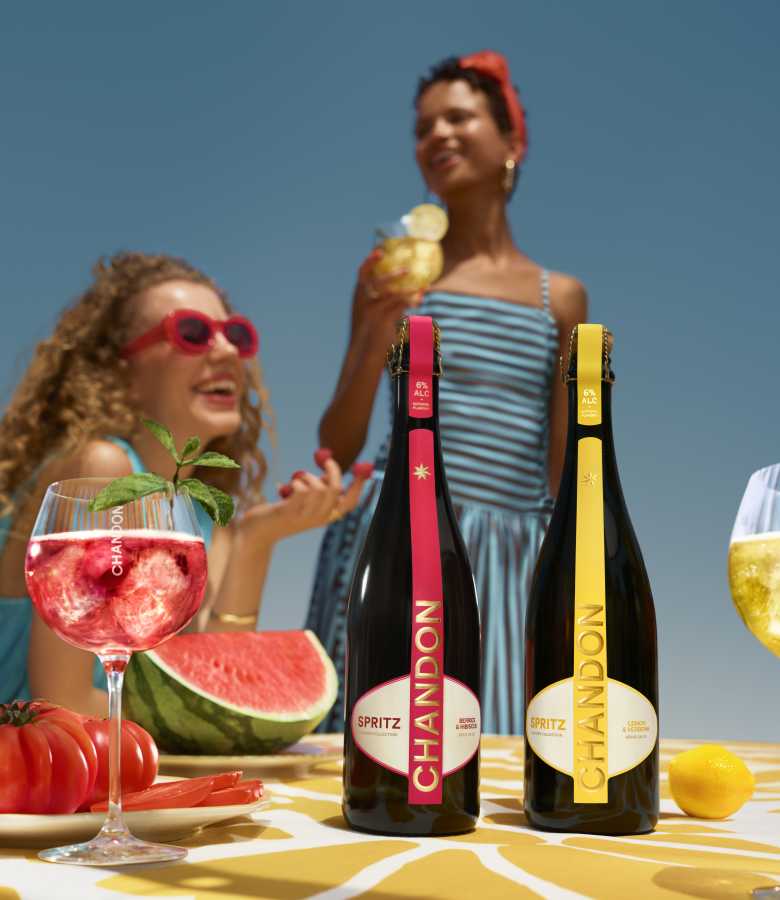 Zwei Flaschen Chandon Spritz (Berries & Hibiscus und Lemon & Verbena) stehen auf einem Tisch, daneben ein Glas roter Chandon Spritz Cocktail, eine Wassermelonenscheibe und frische Beeren. Im verschwommenen Hintergrund sind zwei lächelnde Frauen unter einem klaren blauen Himmel zu sehen.