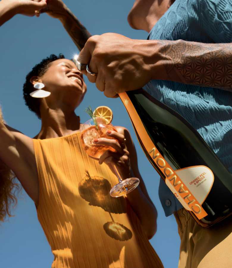 A person's hand holding a bottle of Chandon Spritz Orange Peels & Spices, pouring a drink into a glass held by a smiling woman in a yellow top, both set against a clear blue sky.