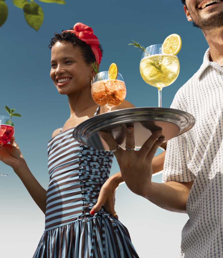 A smiling woman wearing a red headscarf and striped dress, looking upwards, while a person in the foreground holds a silver tray with two Chandon cocktails – one orange and one yellow – garnished with citrus and rosemary.