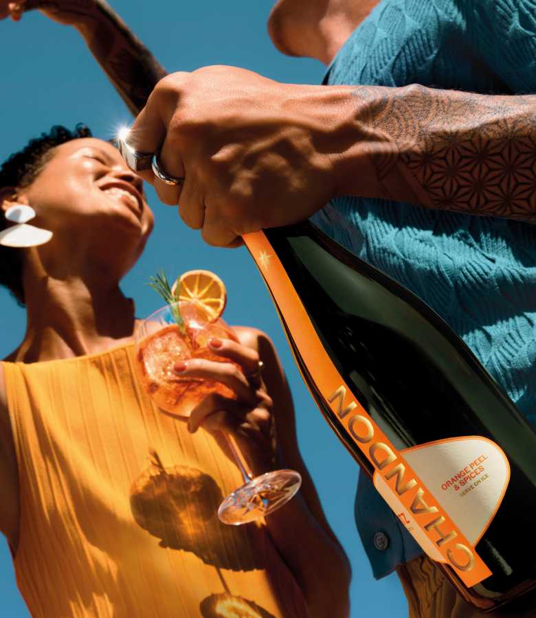 Chandon Spritz Orange cocktail in a glass with orange slice and rosemary, held by a person in an orange top, while another person opens a Chandon sparkling wine bottle. Outdoor summer celebration, refreshing Chandon cocktail recipe, premium sparkling wine experience.