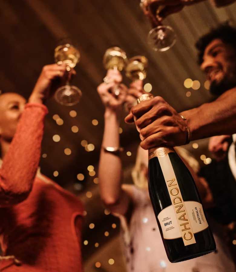 Happy diverse friends toasting with Chandon Brut Australia sparkling wine at a festive evening event with bokeh lights. Celebratory gathering with Chandon wine, group enjoying a sparkling night out, premium event imagery.