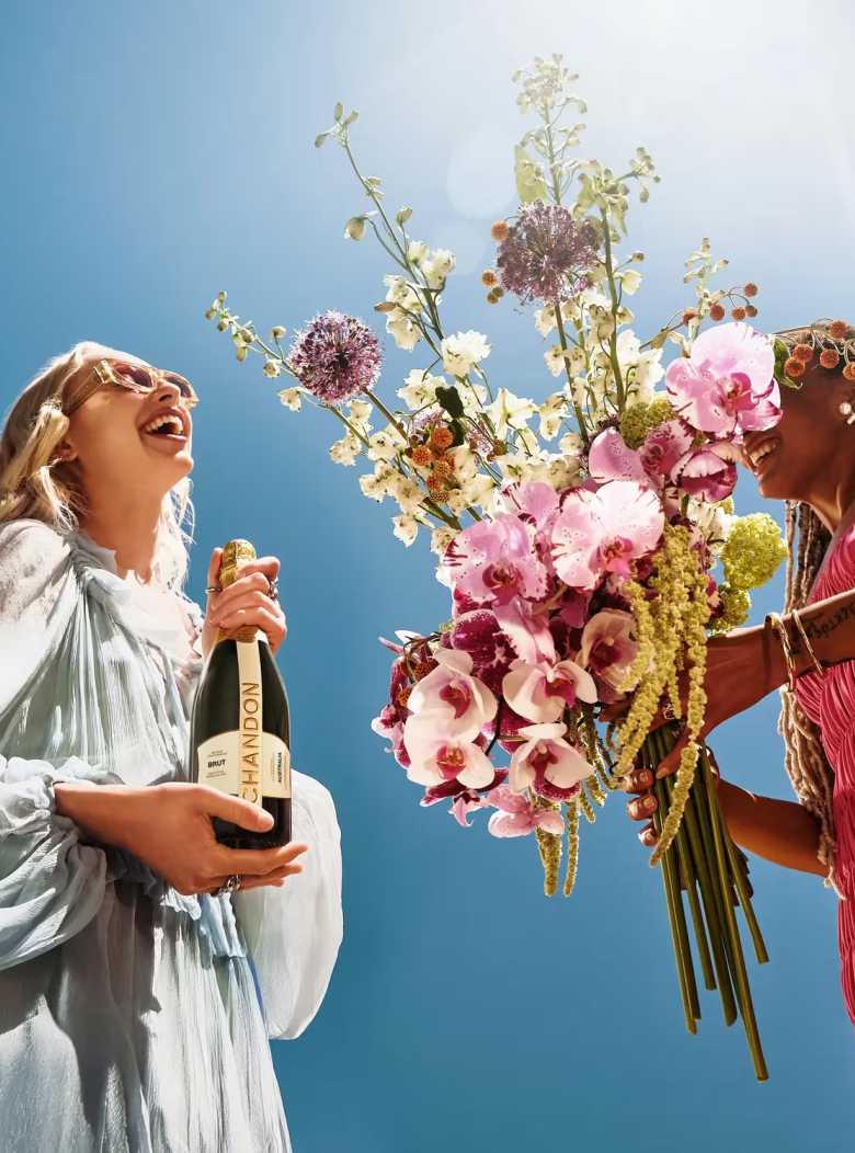Two women celebrating with Chandon Brut Australia sparkling wine and a large, colorful bouquet of flowers under a bright blue sky. A blonde woman in a light blue dress holds the Chandon bottle, smiling, while a woman with dreadlocks in a pink dress presents the elaborate floral arrangement. Ideal for celebratory occasions, gifting, and premium lifestyle branding.