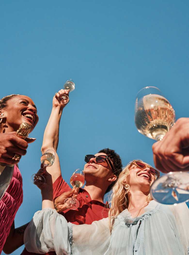 Four diverse friends holding glasses of CHANDON Extra Brut sparkling wine and cocktails, toasting against a clear blue sky, with a CHANDON Extra Brut bottle held by one person, signifying celebration and enjoyment