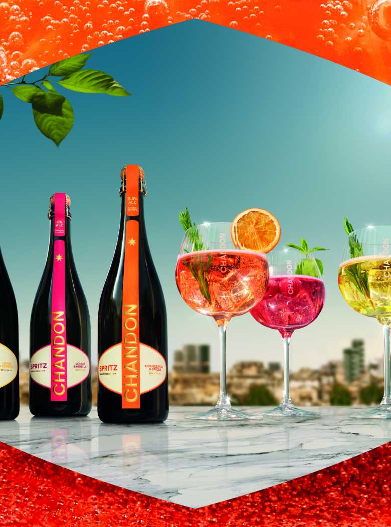 Three bottles of Chandon Spritz (Lemon & Verbena, Berries & Hibiscus, Orange Peels & Spices) displayed alongside three corresponding Chandon cocktails in glasses, garnished with lemon, orange, berries, and mint, all set against a sunny outdoor cityscape. Showcases the full range of refreshing Chandon Spritz flavors perfect for summer gatherings and celebrations.