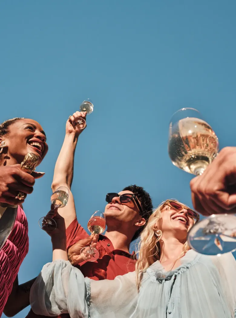 A low-angle shot of a diverse group of four smiling individuals, two women and two men, toasting with glasses of CHANDON sparkling wine against a clear, vibrant blue sky. One woman in the foreground holds a CHANDON bottle, with the brand clearly visible. Their expressions are joyful and celebratory, capturing a moment of shared happiness and festivity.