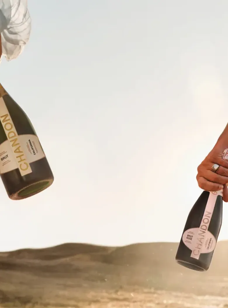 A warm, golden-hour shot of two people from the waist down, walking away from the camera in a sun-drenched, open landscape. One person, on the left, wears a light-colored, flowing garment and carries a full-sized bottle of CHANDON sparkling wine. The person on the right wears darker clothing, has braided hair, and holds a smaller, dark-labeled CHANDON bottle. The setting sun creates a beautiful lens flare, highlighting the relaxed, celebratory atmosphere.