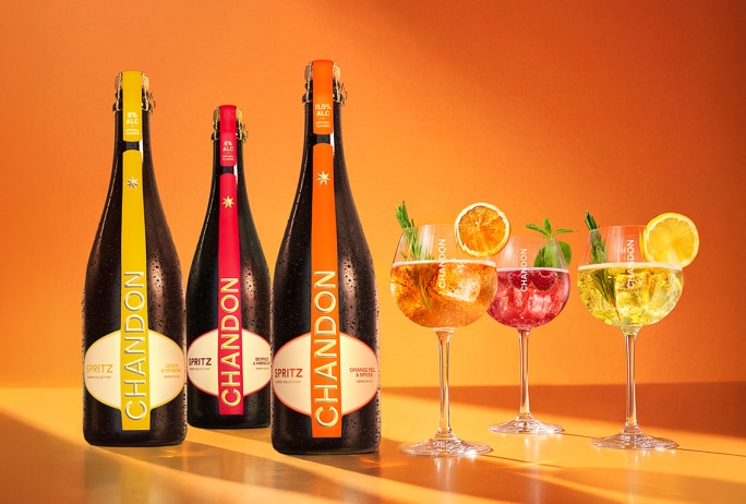 Trois bouteilles de Chandon Spritz de différentes saveurs (jaune pour Lemon & Verbena, rouge pour Berries et Hibiscus, orange pour Orange Peels & Spices) posées sur une surface orange éclatante, avec trois verres de cocktails rafraîchissants, chacun garni de fruits et d'herbes aromatiques. L'image met en valeur la collection Chandon Spritz, idéale pour l'apéritif ou les célébrations estivales.