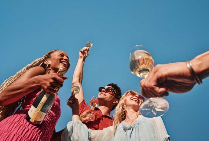A beautifully composed close-up shot of a bottle of CHANDON BRUT AUSTRALIA sparkling wine, prominently displaying its label. To the left, a stemmed glass is being filled with the golden, bubbling wine, with a slight overflow creating a sense of generosity. Another full glass of sparkling wine with elegant bubbles sits to the right, poised for enjoyment. The background is softly blurred, focusing attention on the wine and its celebratory presentation.