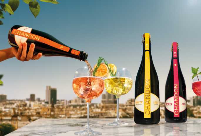 Three bottles of Chandon Spritz (Lemon & Verbena, Berries & Hibiscus, Orange Peels & Spices) displayed alongside three corresponding Chandon cocktails in glasses, garnished with lemon, orange, berries, and mint, all set against a sunny outdoor cityscape. Showcases the full range of refreshing Chandon Spritz flavors perfect for summer gatherings and celebrations.