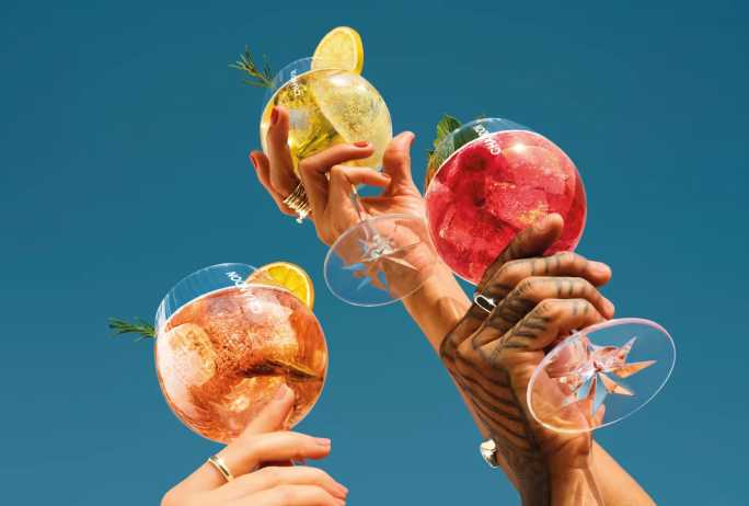 Nahaufnahme von drei Händen, die Chandon Spritz Cocktailgläser in den Sorten Orange, Lemon und Berries halten, garniert mit Zitrusfrüchten, Himbeeren, Rosmarinund Minze, vor einem leuchtend blauen Himmel.