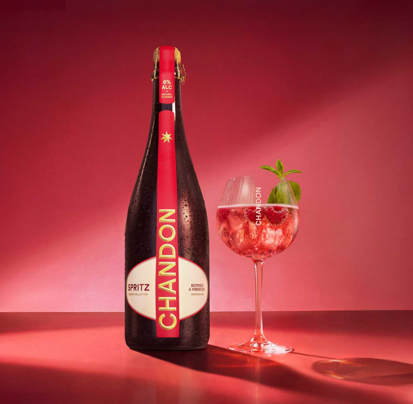 Une bouteille de Chandon Spritz Baies & Hibiscus à côté d'un verre à vin, avec des glaçons, des framboises et des feuilles de menthe dispersés sur un fond rouge vif.