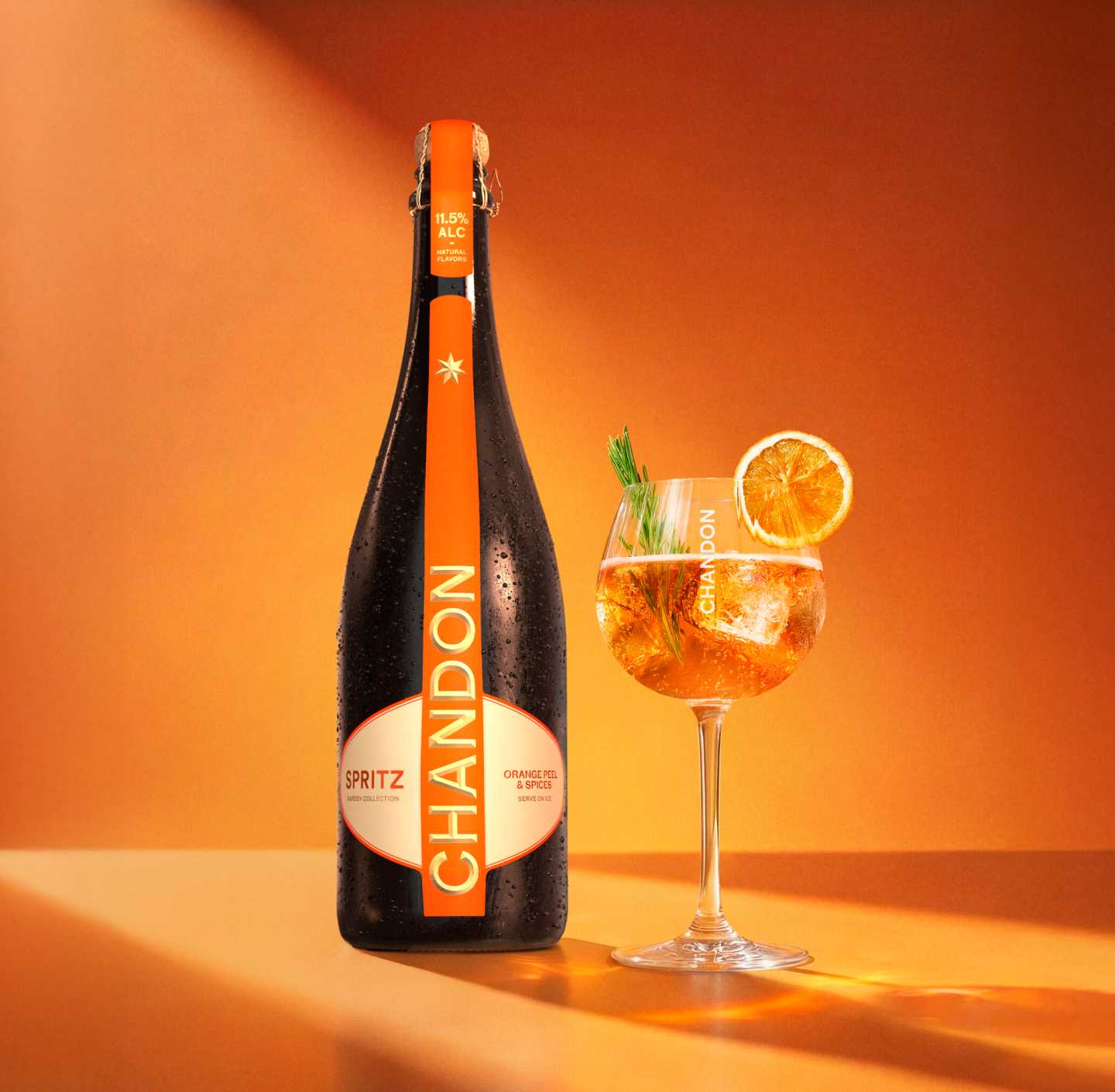 Bouteille de Chandon Spritz, tranche d'orange séchée, brin de romarin et glaçons disposés sur un fond orange à côté du contour embossé d'un verre à vin.