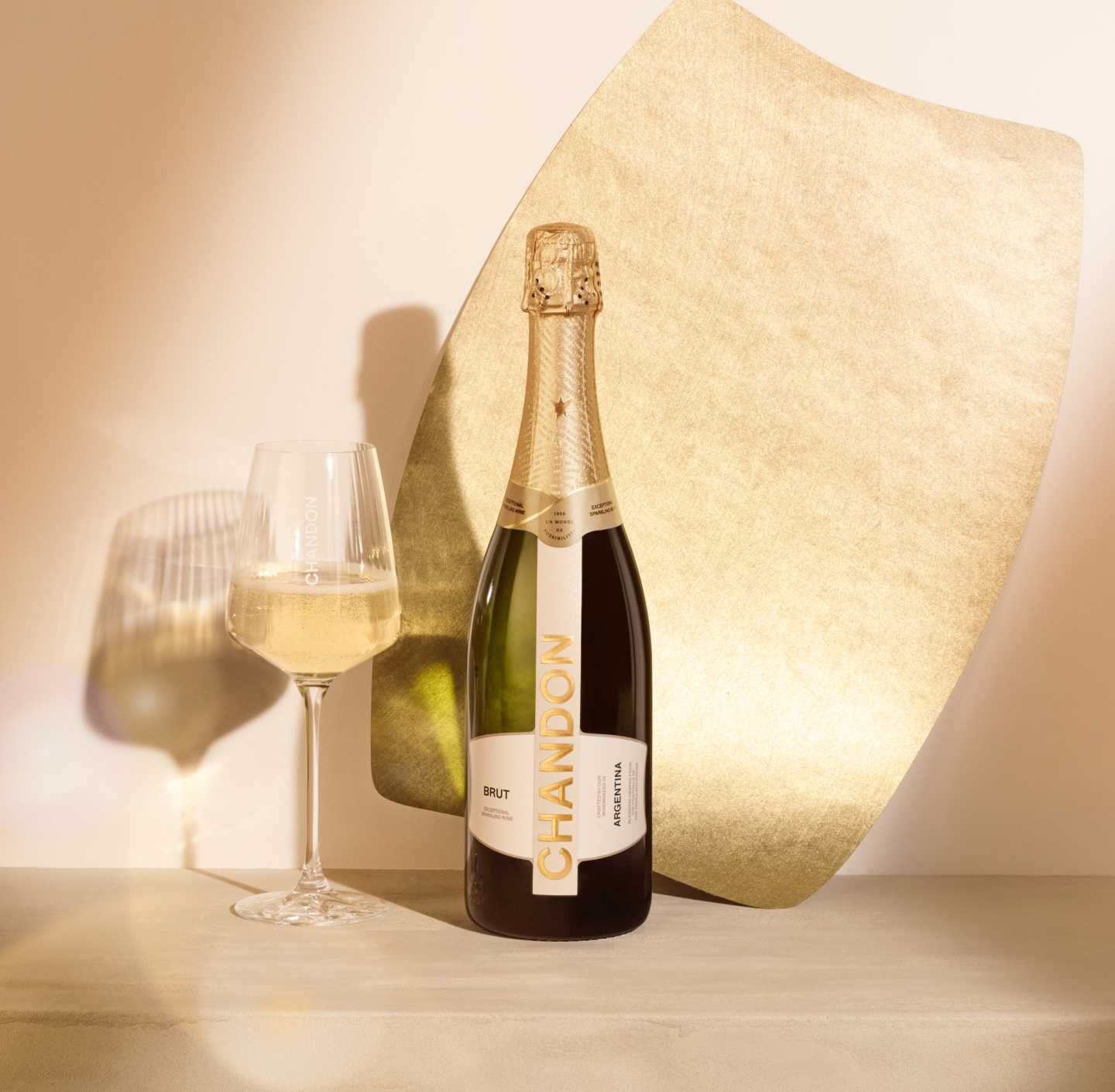 A bottle of CHANDON Brut sparkling wine from Argentina, with a gold and white label, next to an elegant wine glass filled with clear, sparkling white wine, arranged against a modern, minimalist background of an abstract, textured gold panel.