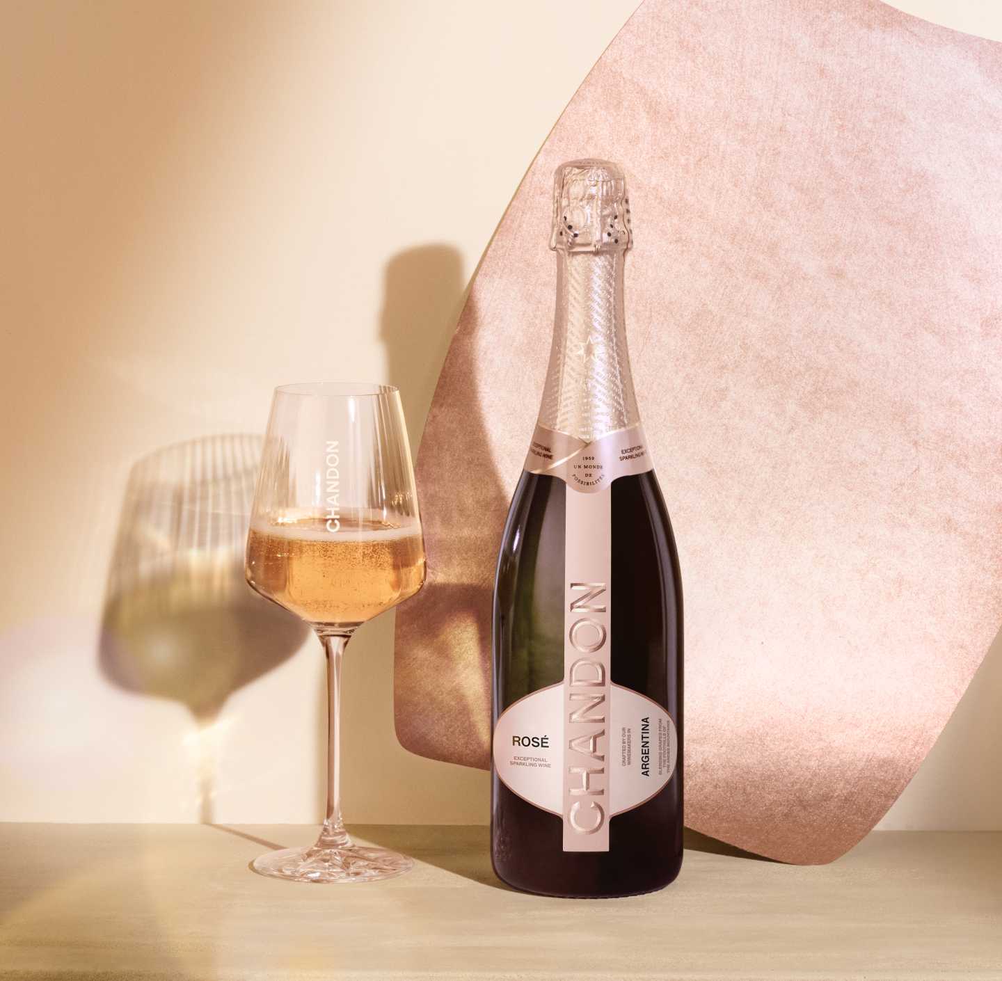 A bottle of CHANDON Rosé sparkling wine from Argentina, with a rose-colored label, next to an elegant wine glass filled with pink sparkling wine, arranged against a modern, minimalist background of an abstract, textured rose gold panel.