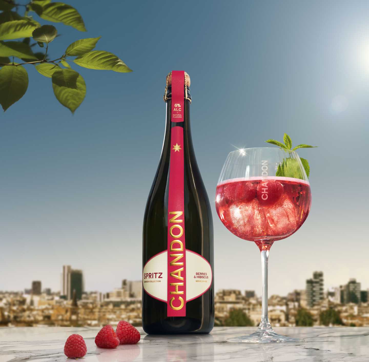 Eine Flasche Chandon Spritz Berries & Hibiscus neben einem Glas Chandon Spritz garniert mit frischen Himbeeren und einem Minzblatt, vor dem Hintergrund einer sonnigen Stadtlandschaft und unter blauem Himmel.