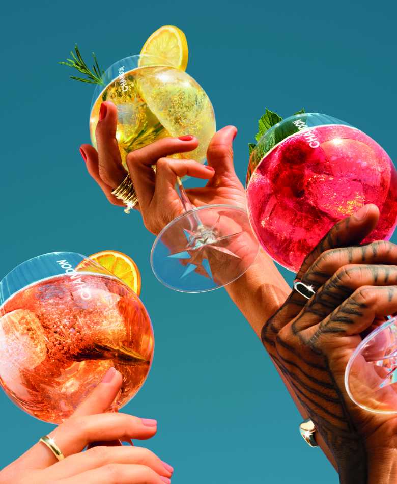 Nahaufnahme von drei Händen, die Chandon Spritz Orange, Lemon und Berries Cocktailgläser halten, garniert mit Zitrusfrüchten und Rosmarin, vor einem leuchtend blauen Himmel