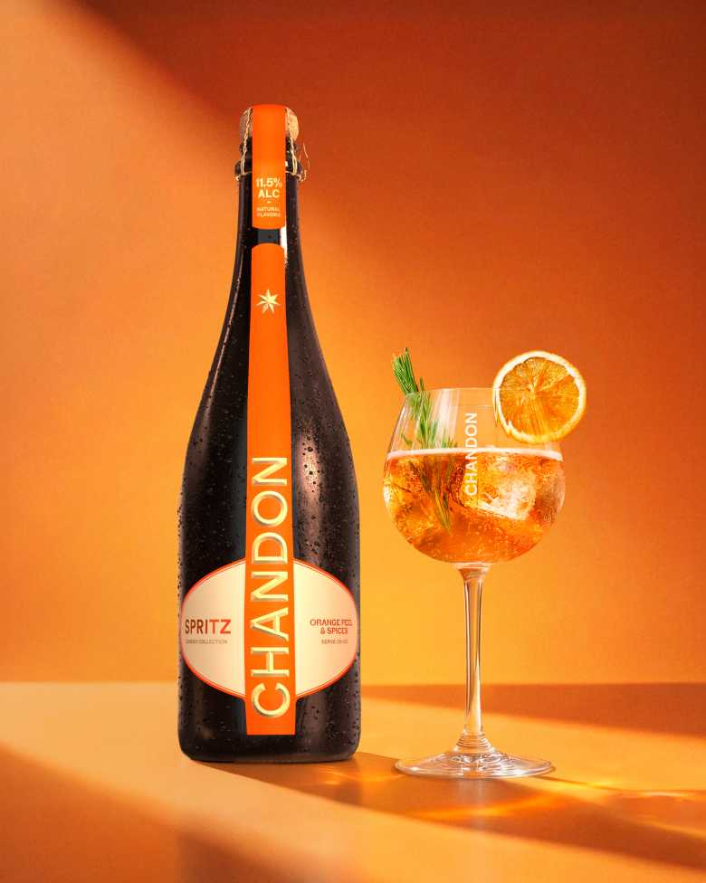 Eine einzelne Flasche Chandon Spritz Orange Peels & Spices neben einem Glas Chandon Spritz, garniert mit einer getrockneten Orangenscheibe und einem Rosmarinzweig, alles auf einem orangefarbenen Hintergrund mit Farbverlauf.