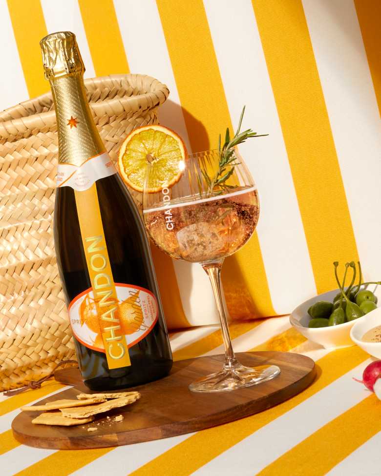 Bottle of CHANDON Garden Spritz next to a cocktail glass with a dried orange slice and rosemary, placed on a wooden board with crackers, a wicker basket, and a yellow and white striped background.