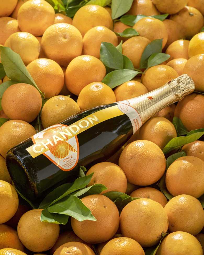 Bottle of CHANDON Garden Spritz nestled among a vibrant pile of fresh, ripe oranges with green leaves, emphasizing the natural fruit ingredients.