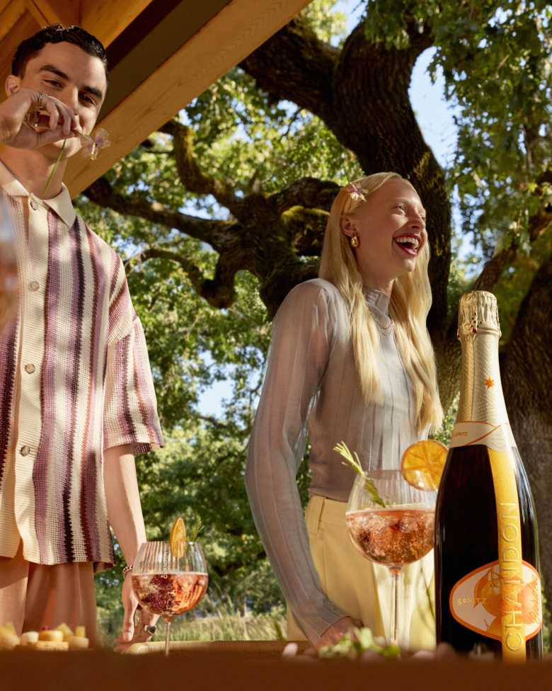 Two happy people enjoying CHANDON Garden Spritz cocktails outdoors, with a bottle of CHANDON on a wooden surface in the foreground and a large green tree in the background, suggesting a relaxed social gathering.