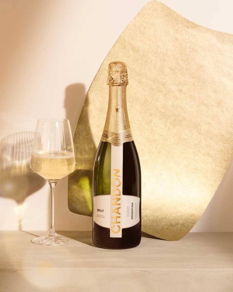A bottle of CHANDON Brut sparkling wine from Argentina, with a gold and white label, next to an elegant wine glass filled with clear, sparkling white wine, arranged against a modern, minimalist background of an abstract, textured gold panel.