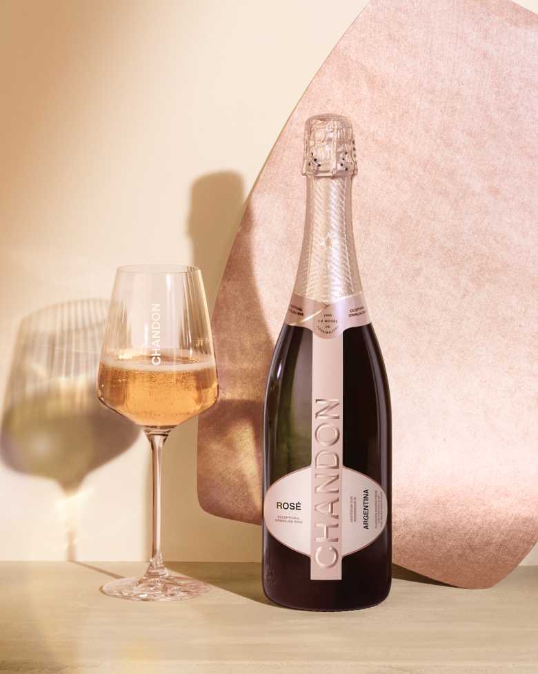 A bottle of CHANDON Rosé sparkling wine from Argentina, with a rose-colored label, next to an elegant wine glass filled with pink sparkling wine, arranged against a modern, minimalist background of an abstract, textured rose gold panel.