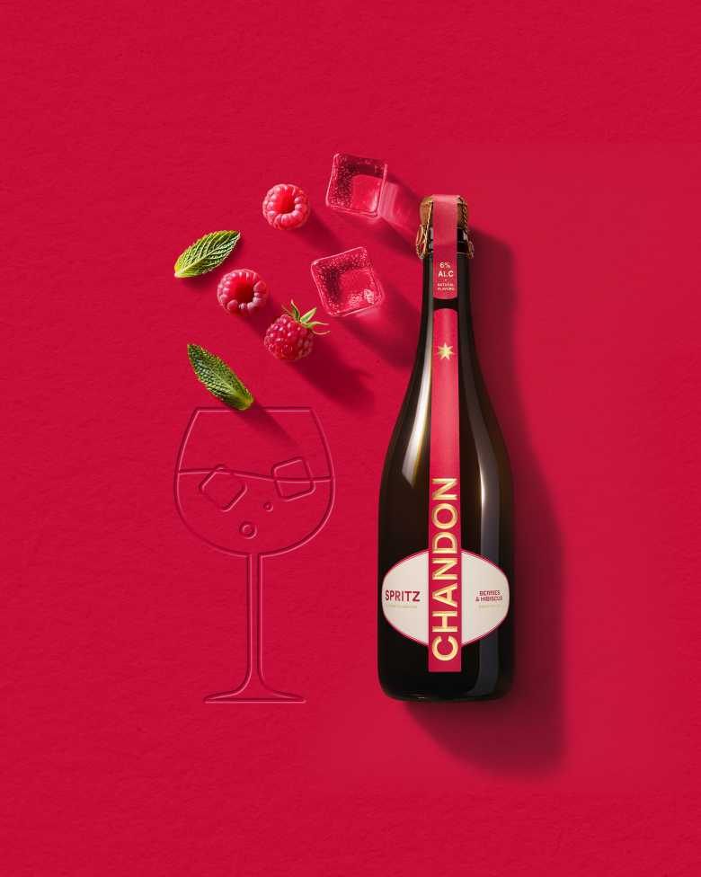 Eine Flasche Chandon Spritz Berries & Hibiscus neben einer Weinglaskontur, mit Eiswürfeln, Himbeeren und Minzblättern auf einem kräftig roten Hintergrund.