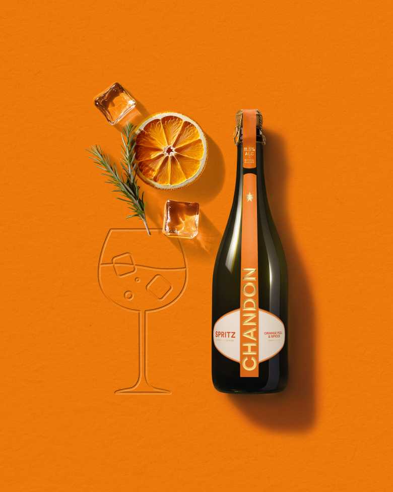 Chandon Spritz bottle, dried orange slice, rosemary sprig, and ice cubes arranged on an orange background next to an embossed outline of a wine glass