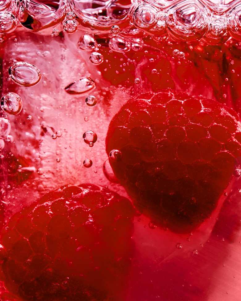 Nahaufnahme von zwei frischen Himbeeren, die in einer sprudelnden, roten Flüssigkeit versinken.