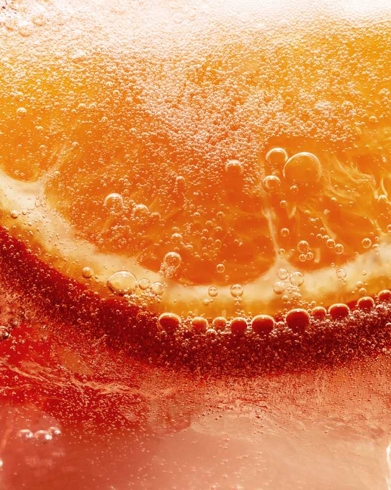 Nahaufnahme einer Orangenscheibe, die in einer sprudelnden, orangefarbenen Flüssigkeit versunken ist.