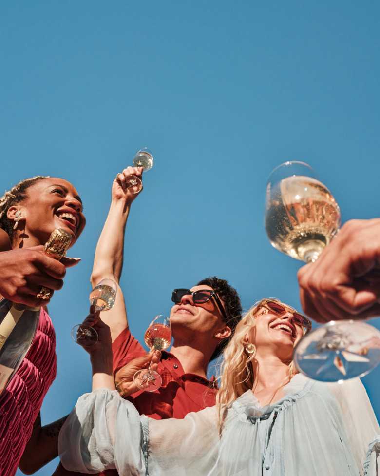 Four diverse friends holding glasses of CHANDON Extra Brut sparkling wine and cocktails, toasting against a clear blue sky, with a CHANDON Extra Brut bottle held by one person, signifying celebration and enjoyment