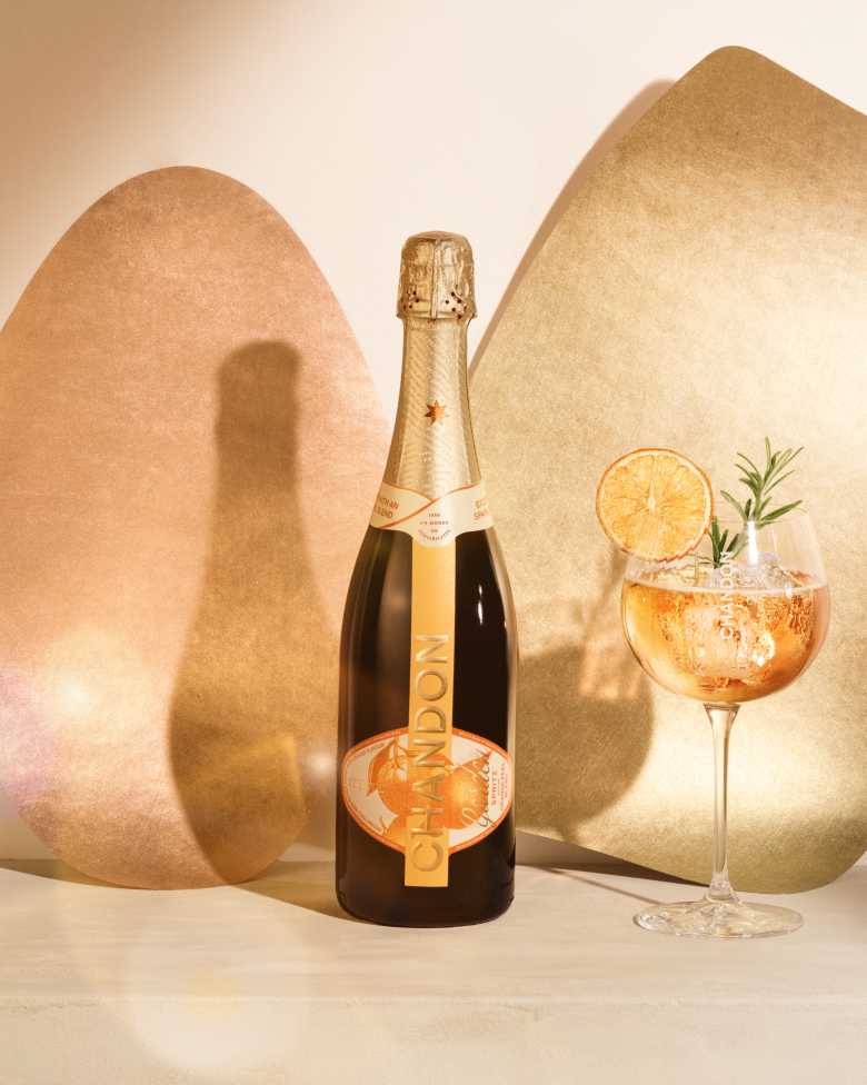 A bottle of CHANDON Garden Spritz and a glass of the spritz cocktail garnished with a dried orange slice and rosemary, against a modern, minimalist background of two abstract, textured panels in rose gold and gold hues.