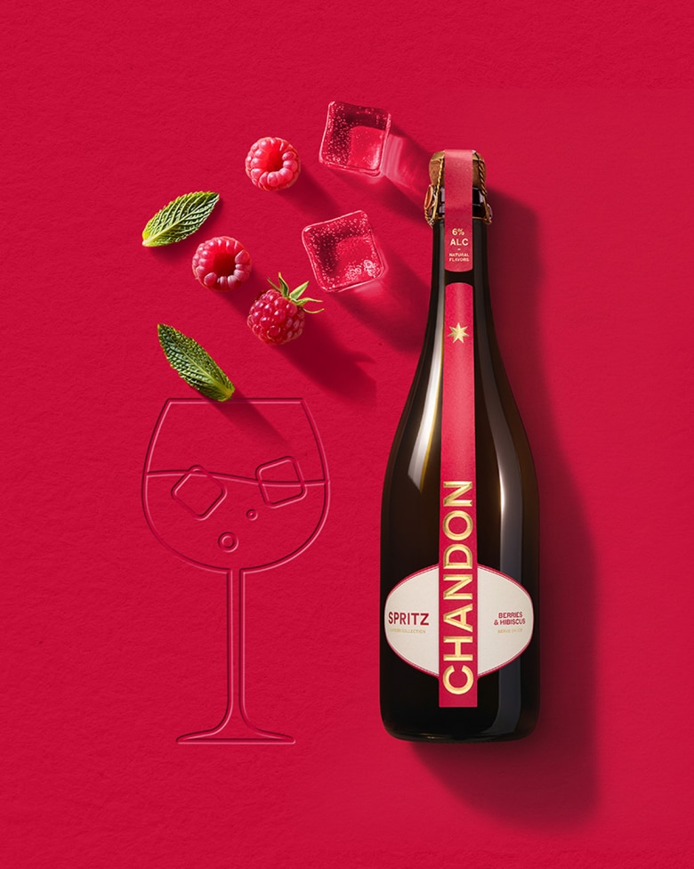 Une bouteille de Chandon Spritz Baies & Hibiscus à côté du contour d'un verre à vin, avec des glaçons, des framboises et des feuilles de menthe dispersés sur un fond rouge vif.