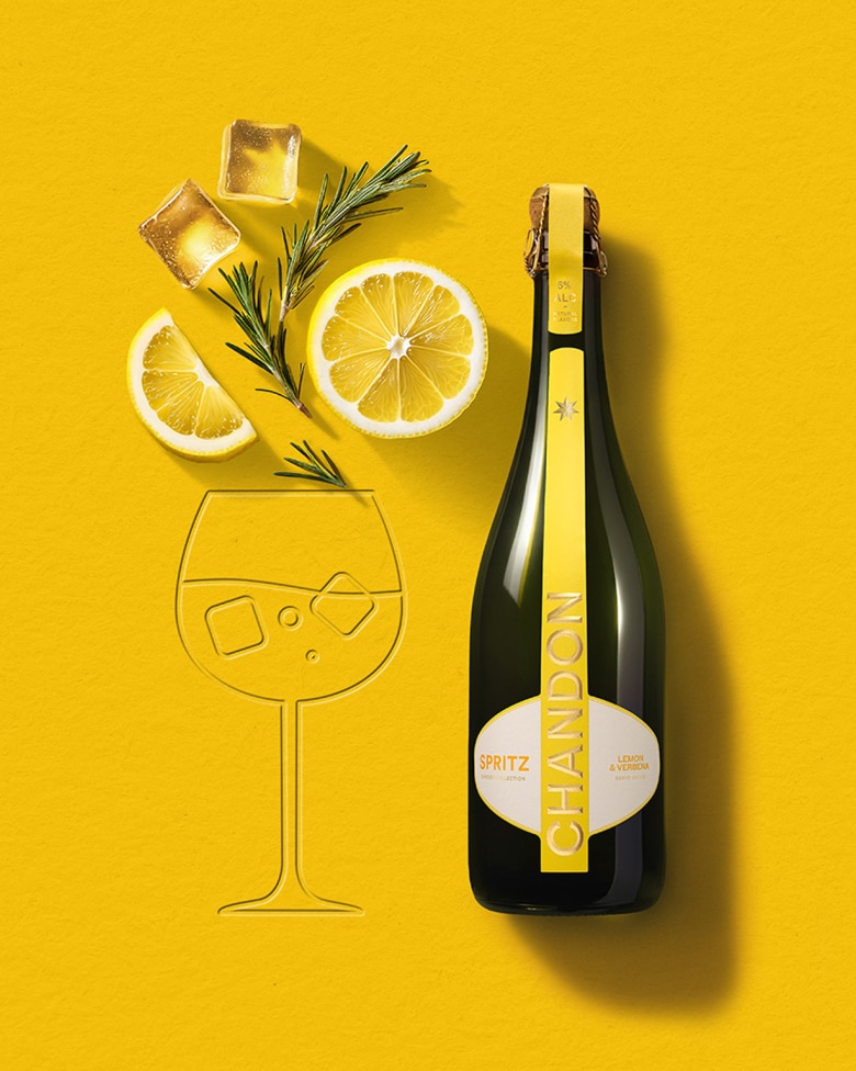 Une bouteille de Chandon Spritz Citron & Verveine à côté du contour d'un verre à vin, avec des glaçons, une tranche de citron et un brin de romarin disposés sur un fond jaune vif.